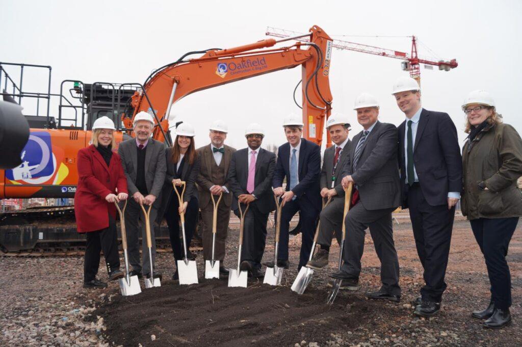 10 people standing in a field, facing towards the camera with a smile. They are wearing suits and hardhats and are all stood around a pile of dirt with their spades in the ground. Behind them is a large orange digger